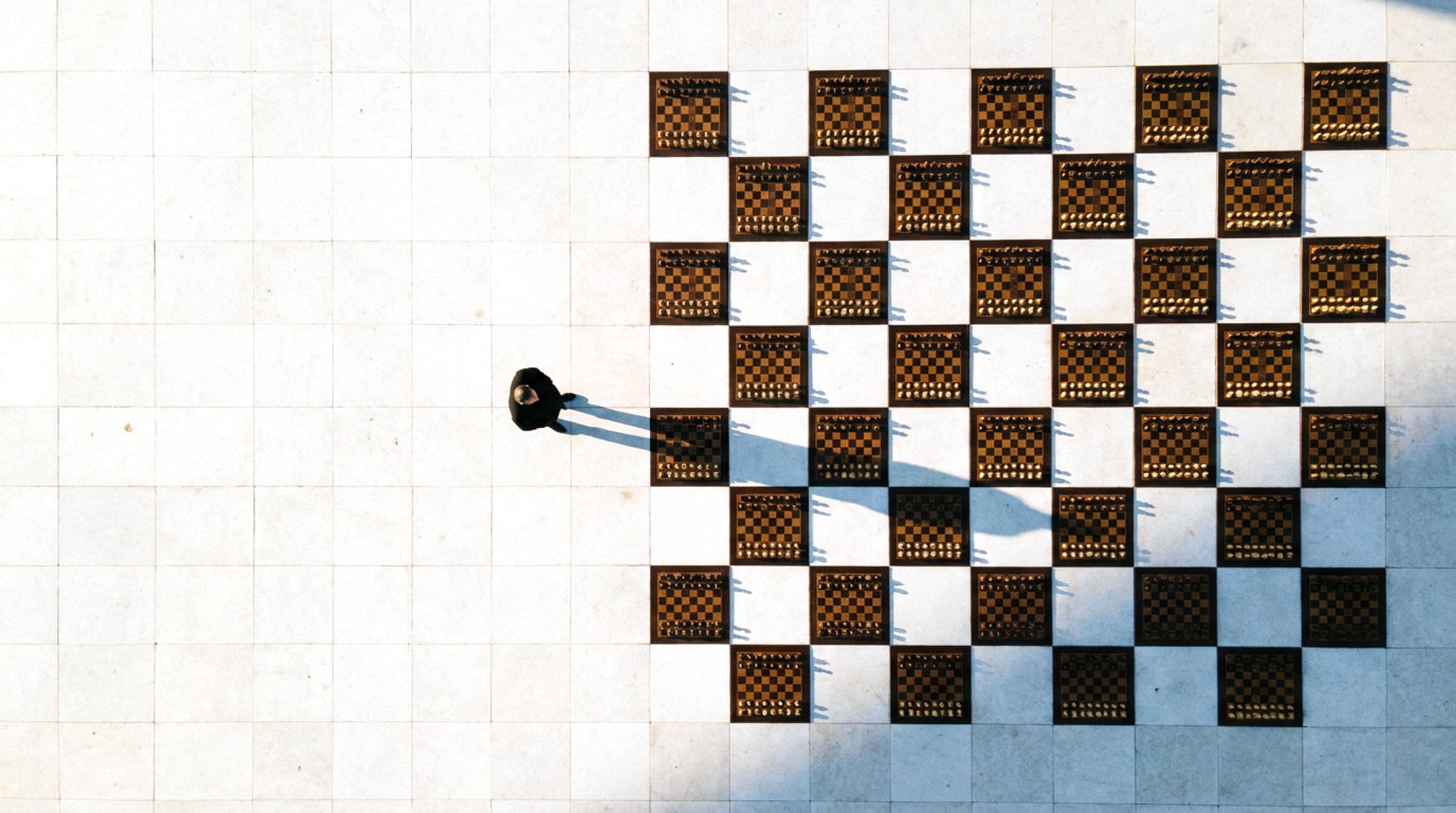 Seen from above: dozens of chess boards laid out on a pale stone plaza in a giant chequerboard, every game in play, with one person standing among them.